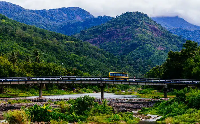 È un'immagine che mostra un autobus giallo su un ponte che attraversa un fiume, con montagne sullo sfondo, lungo una strada di campagna in Thailandia. Il paesaggio è molto verde e rigoglioso
